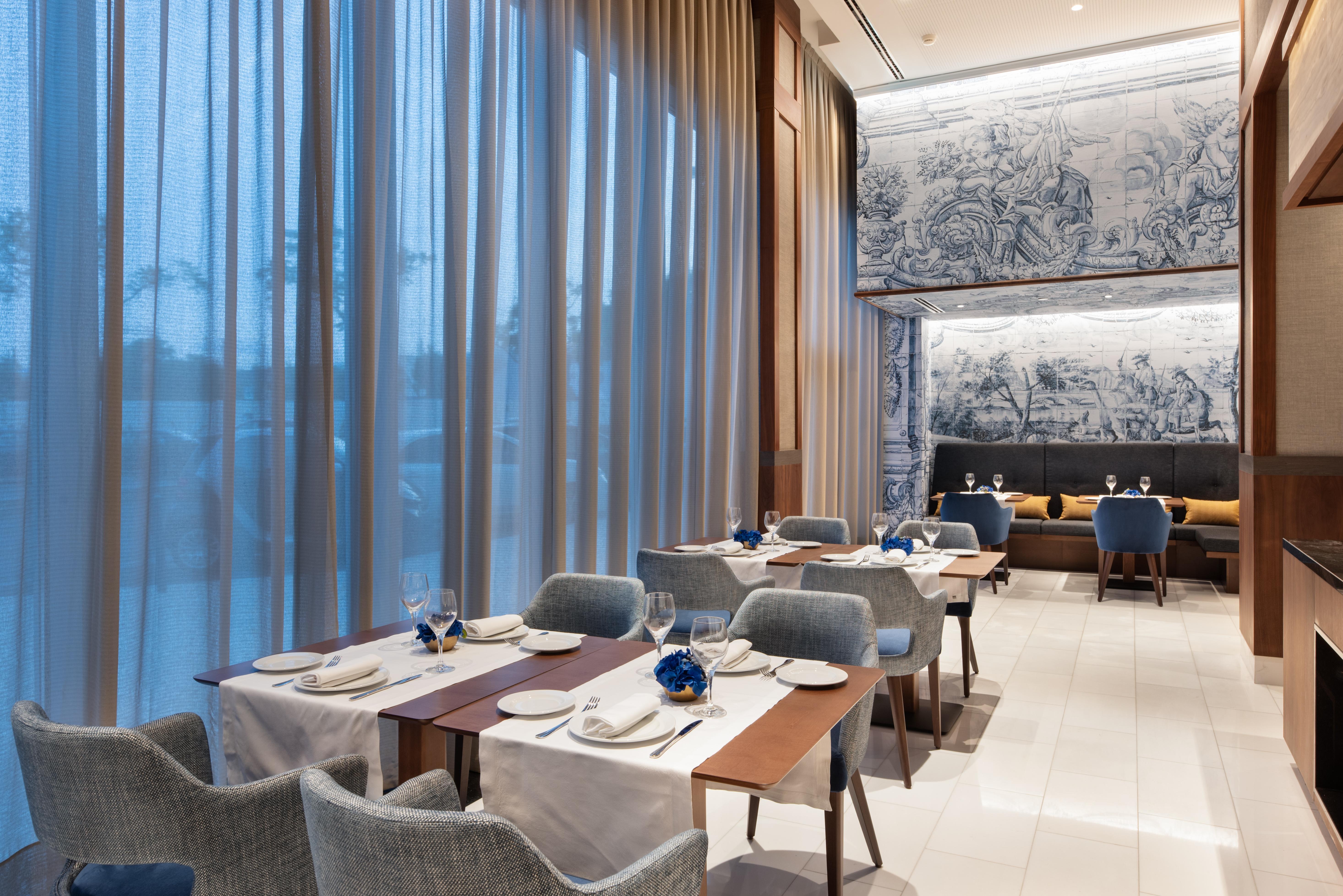 Eurostars Universal Lisboa Hotel Exterior photo The photo shows a modern dining area in a restaurant. There are several tables set with white tablecloths, plates, glasses, and decorative elements. The chairs are upholstered in a soft blue fabric. Large windows draped with sheer curtains allow natu