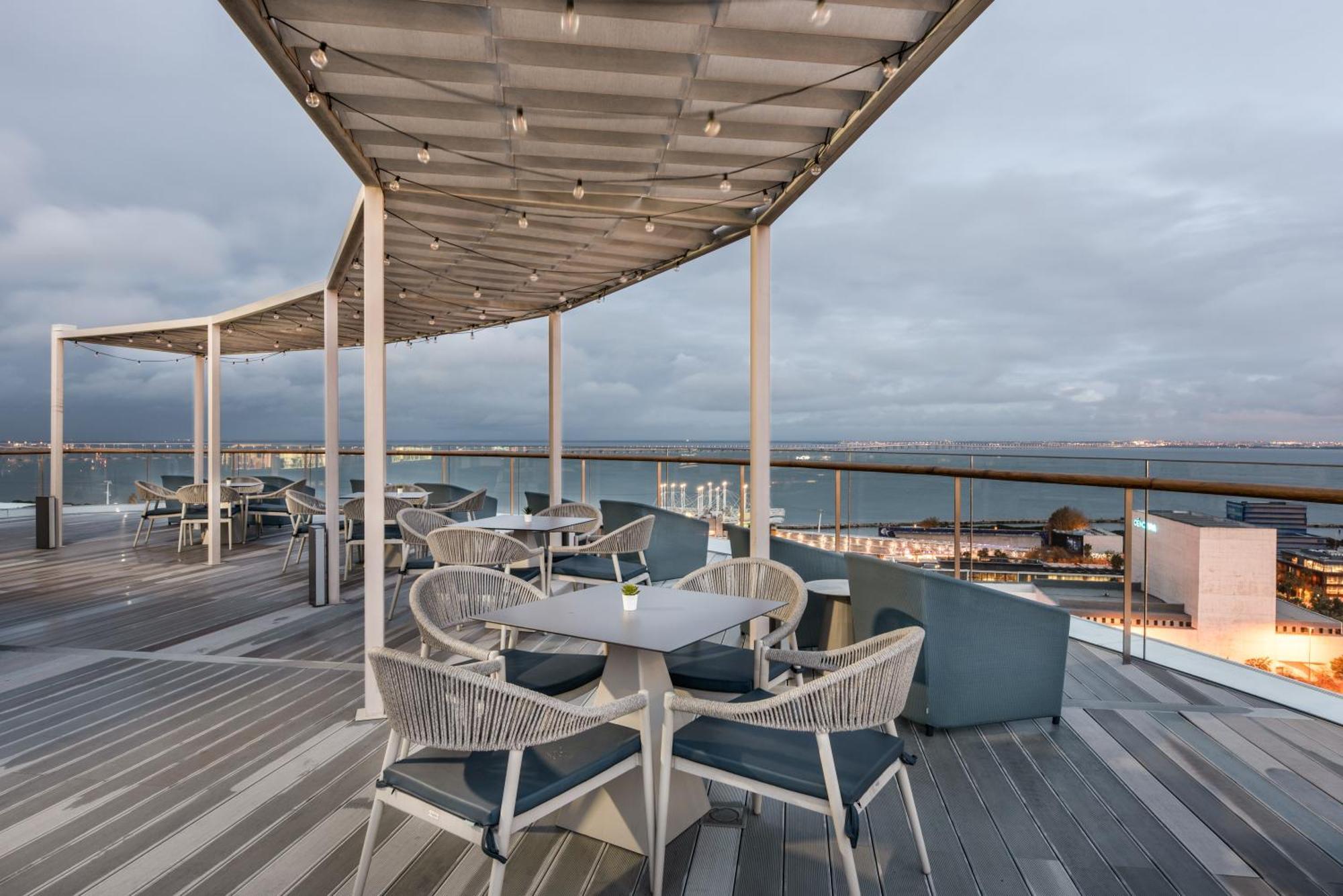 Eurostars Universal Lisboa Hotel Exterior photo The photo shows a scenic outdoor dining area with modern furniture. There are several round tables and chairs arranged on a wooden deck. Some of the tables have blue cushions, and the area is covered by a sleek, curved canopy that is adorned with str