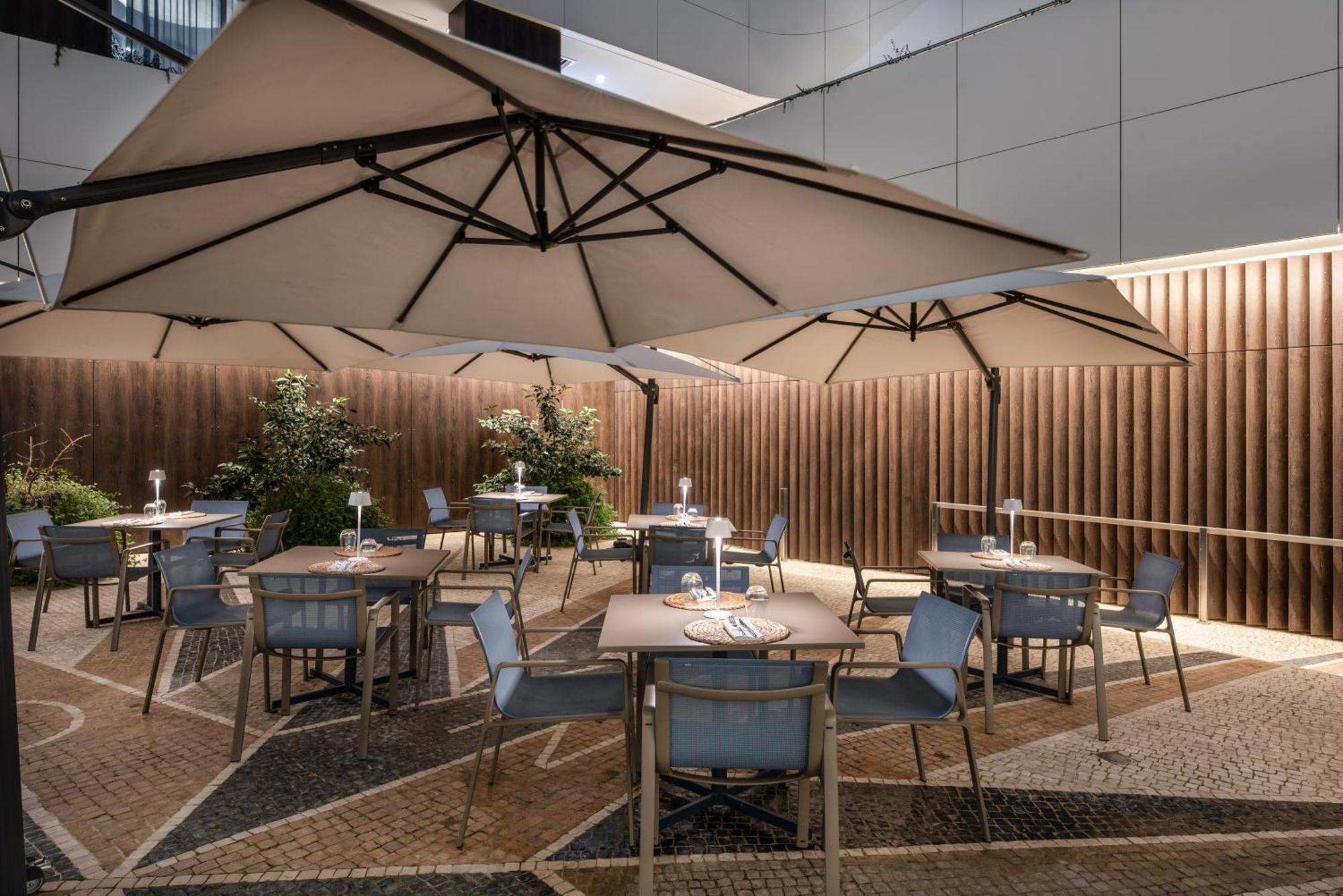 Eurostars Universal Lisboa Hotel Exterior photo The photo depicts an outdoor dining area featuring several tables and chairs arranged under large umbrellas. The setting is likely part of a restaurant or café, with a nicely designed stone or tiled floor. There are plants in the background, providin